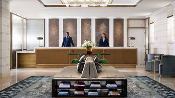 Zona de recepción con mostrador, dos recepcionistas, un centro de mesa decorativo y una estantería con libros.