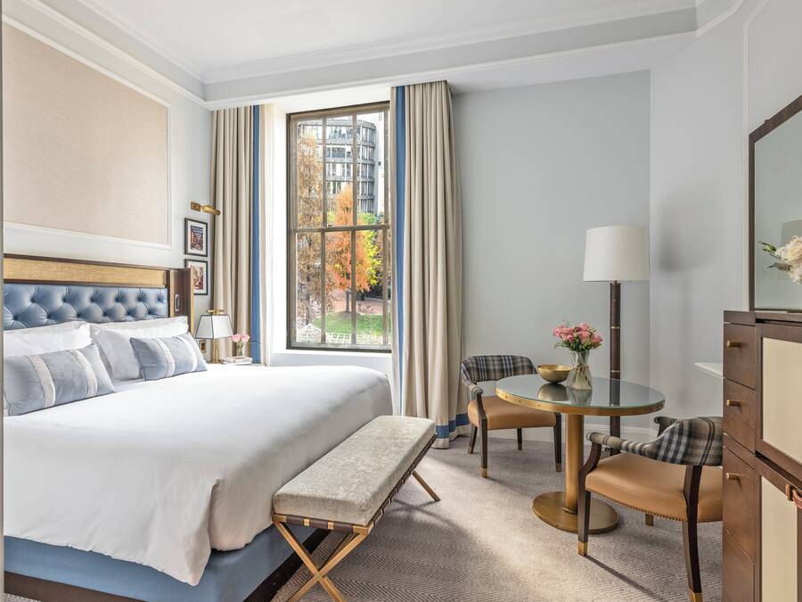 A hotel room with a large bed, a bench, a round table with chairs, a desk, and a mirror.