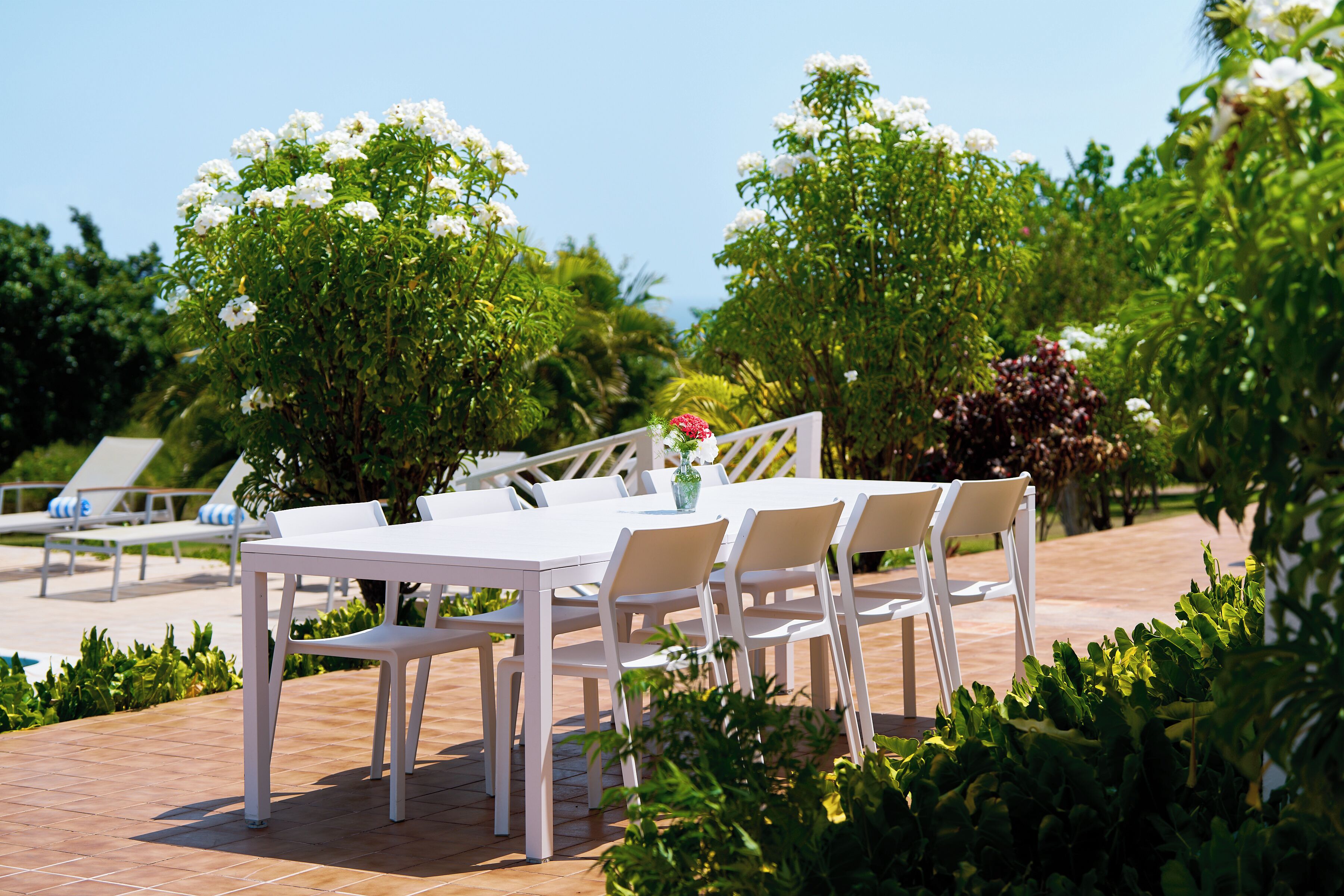 Un espacio exterior para comer con una mesa y sillas blancas, rodeado de abundante follaje y plantas con flores blancas.