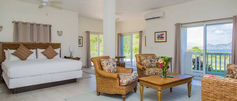 A spacious bedroom with a large bed, two wicker armchairs, a wooden coffee table, and a balcony with a view.