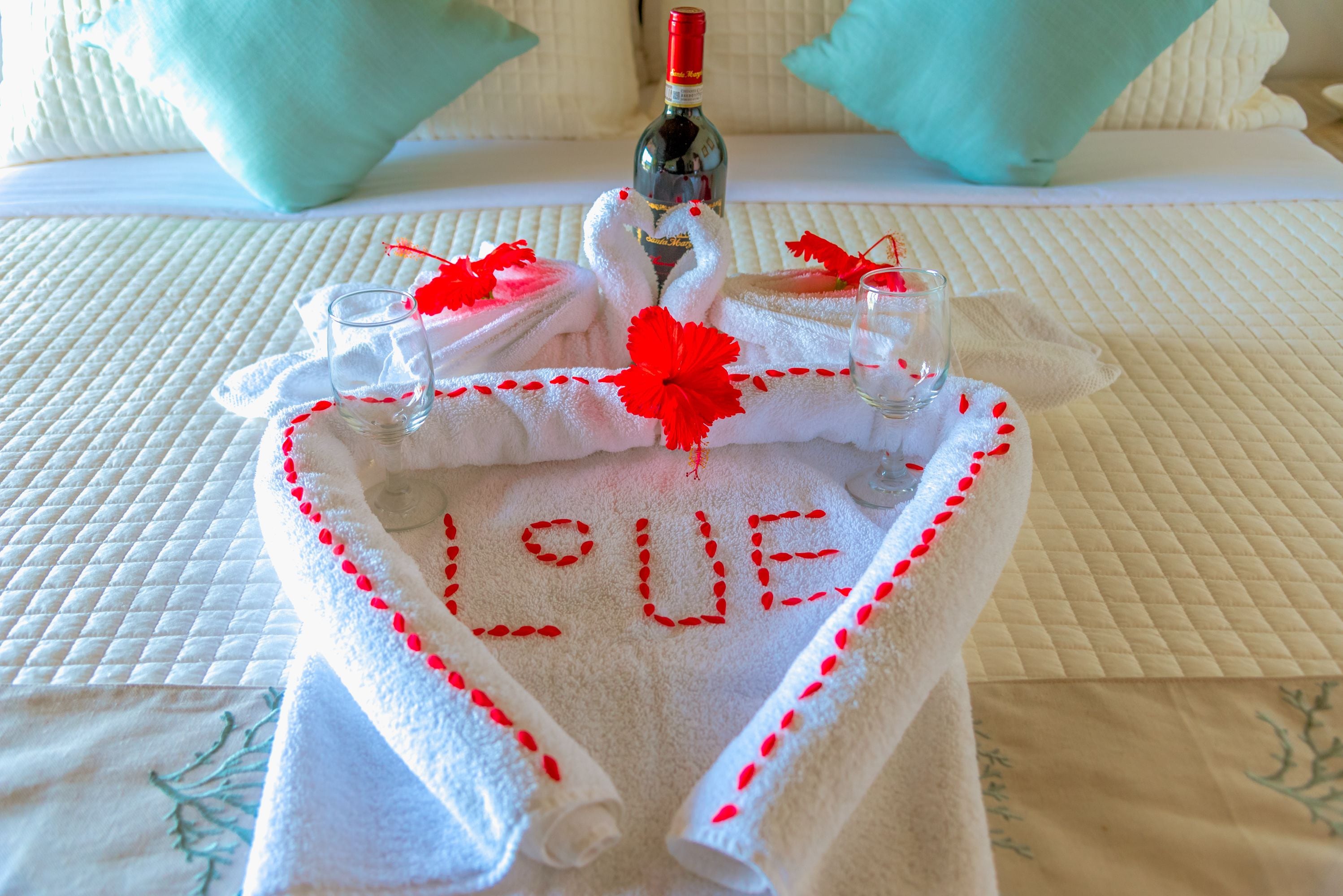 Una cama con una colcha blanca acolchada, una toalla con la palabra 'LOVE' bordada en rojo, dos vasos de agua y una botella de vino.