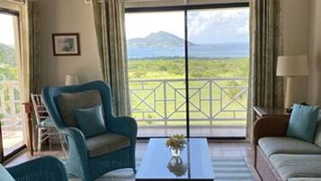 Ein Zimmer mit Holzboden, einem großen Fenster mit Blick auf Berge und Grünflächen, zwei blaue Sessel, ein Couchtisch aus Holz mit einer Vase und ein beiges Sofa.