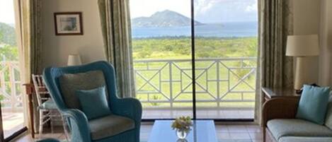 Una habitación con techo de madera, un ventanal con vistas a una montaña y vegetación, dos sillones azules, una mesa de centro de madera con un jarrón, y un sofá color beige.