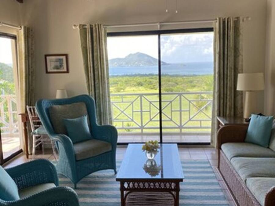 Ein Zimmer mit Holzboden, einem großen Fenster mit Blick auf Berge und Grünflächen, zwei blaue Sessel, ein Couchtisch aus Holz mit einer Vase und ein beiges Sofa.