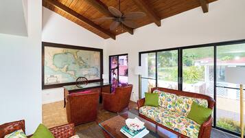 A spacious living area with a wooden ceiling, a large map on the wall, wicker furniture, and large windows offering a view of the outdoors.