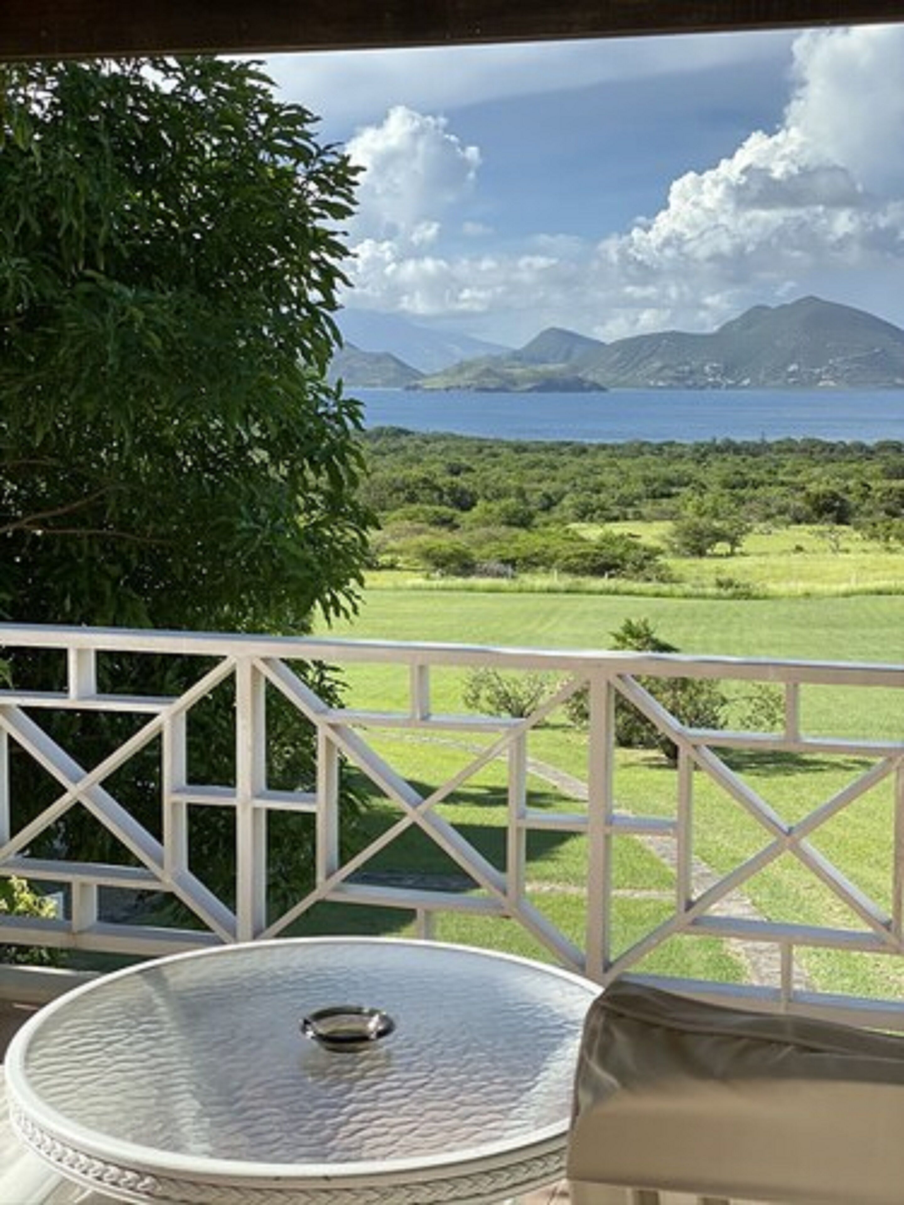 A balcony with a glass table and white railing, overlooking a lush green landscape and a body of water with mountains in the distance.