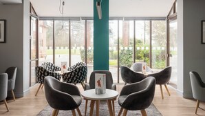 A modern indoor seating area with round tables and chairs, large windows with a view of greenery and buildings, and a teal pillar. - Holiday Inn Northampton by IHG (Northampton)