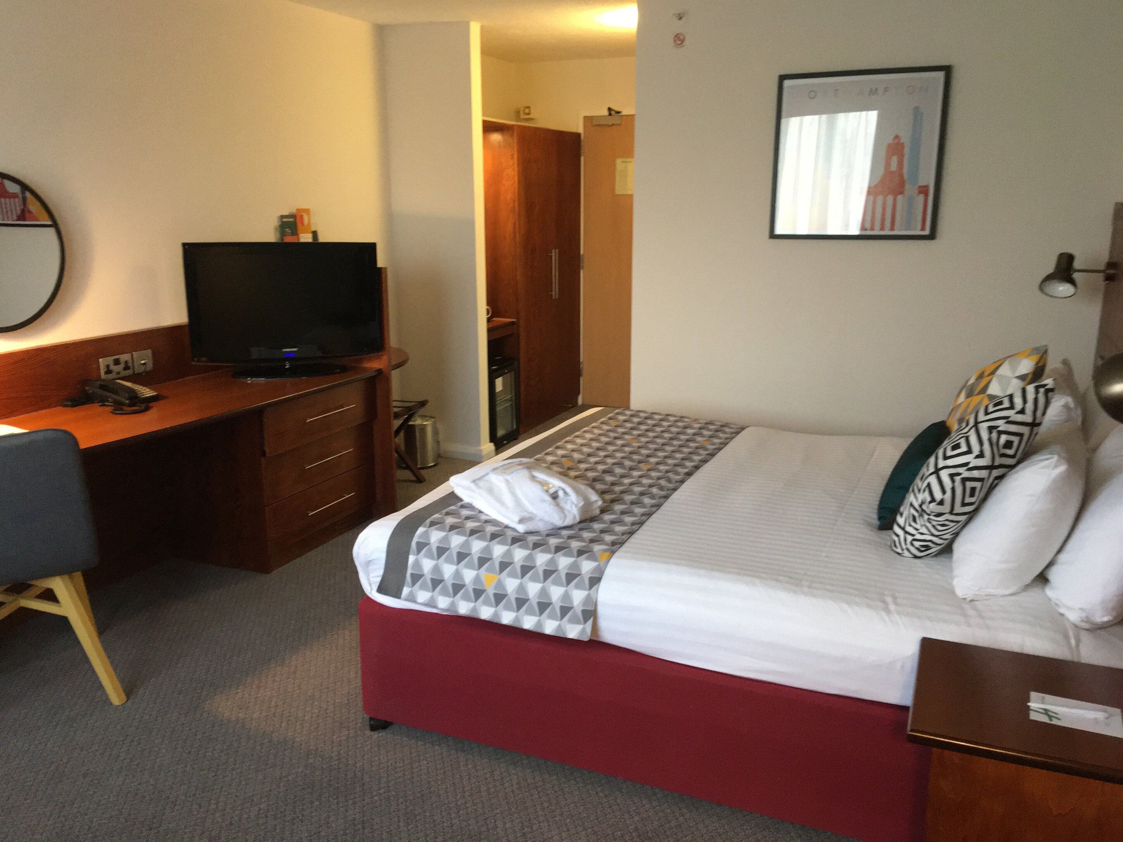 A hotel room with a bed, a desk with a television, a wardrobe, and a framed picture on the wall.