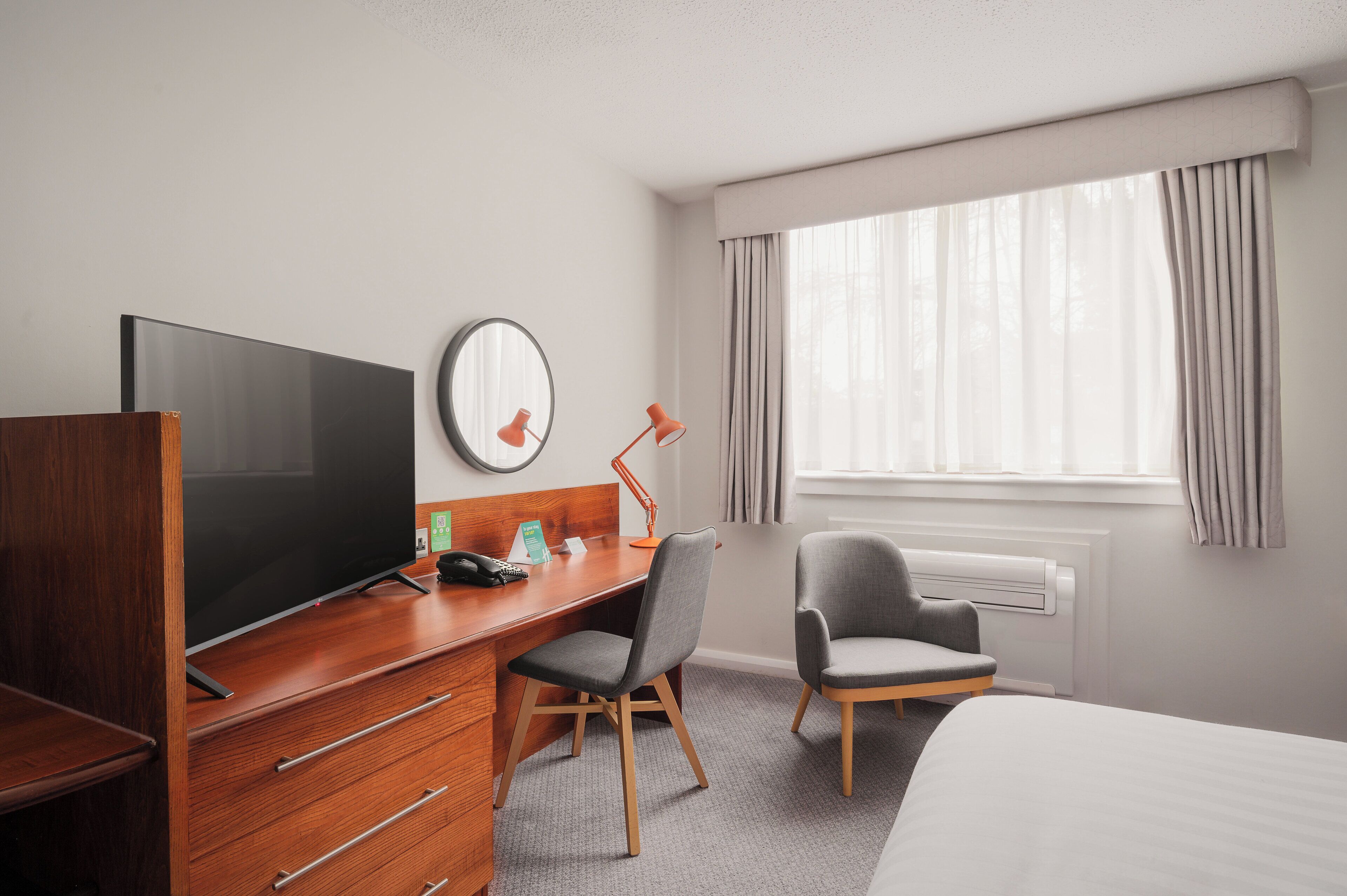 A hotel room with a wooden desk, a television, a chair, a bed, and a window with curtains.