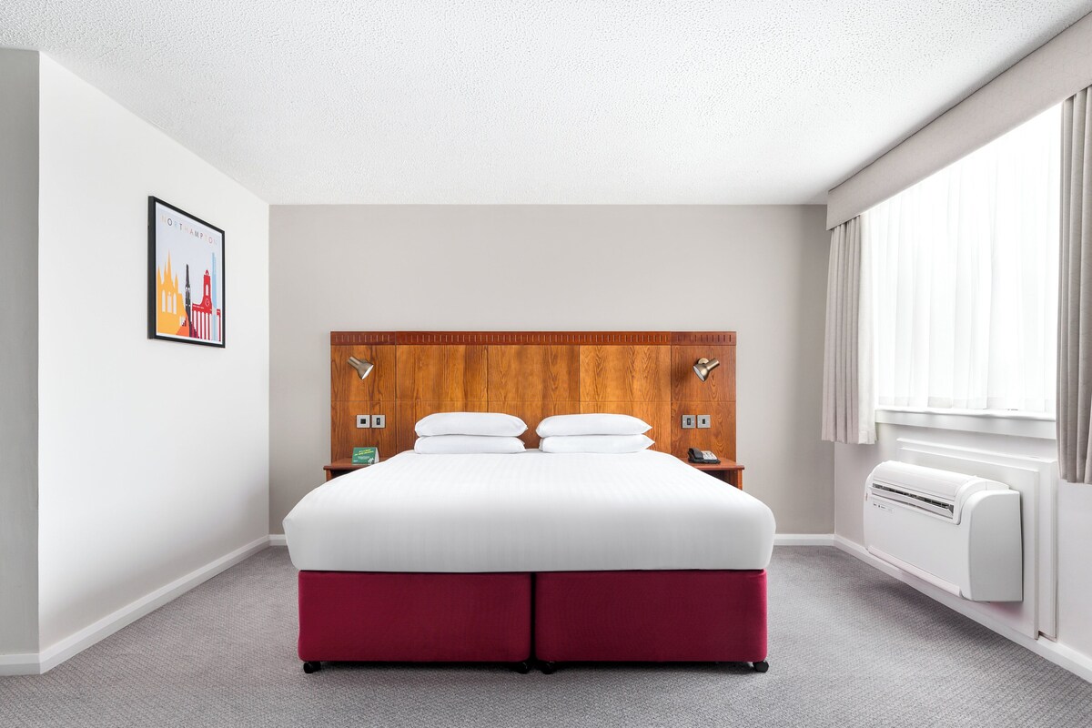 Una habitación de hotel con una cama grande, un cabecero de madera, aire acondicionado de pared y una ventana con cortinas.