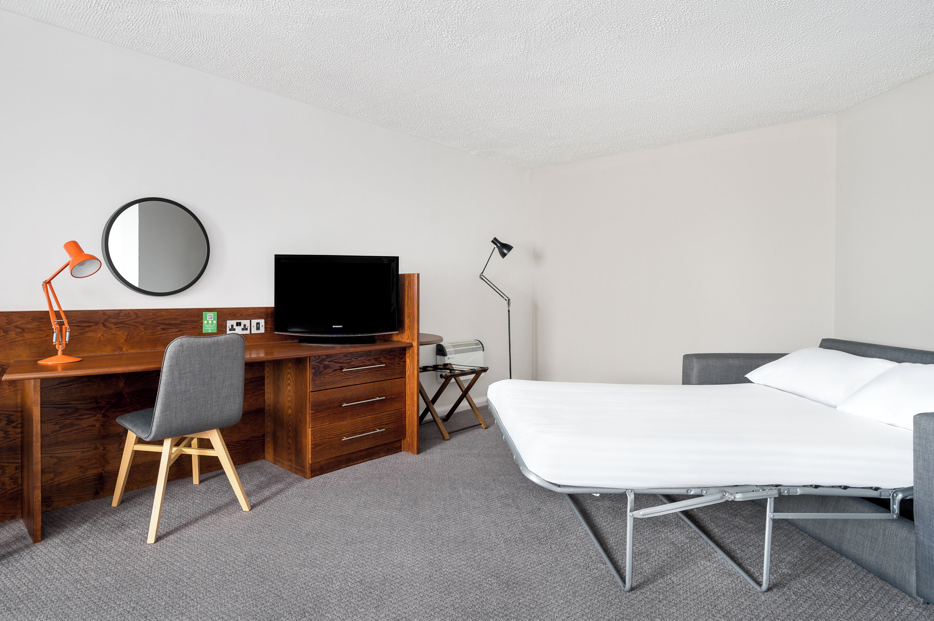 A hotel room with a wooden desk, a television, a bed, and a bedside table.