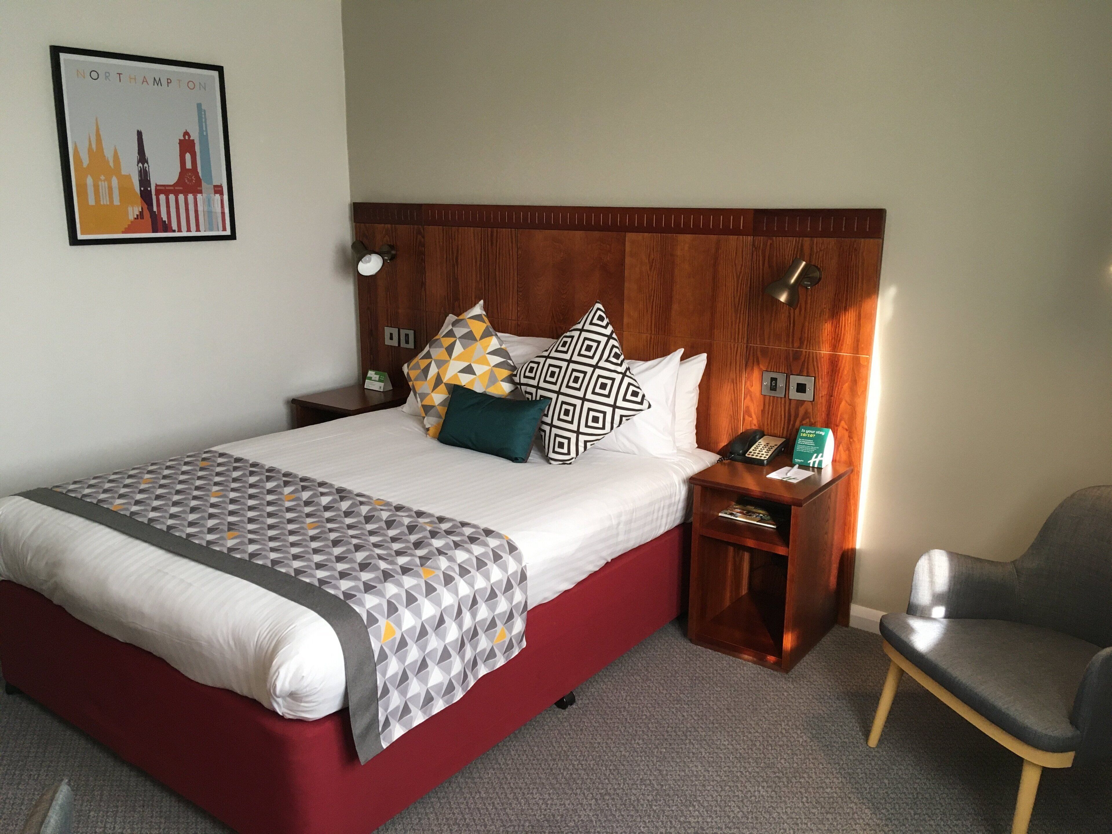 A hotel room with a large bed, a wooden headboard, a bedside table with a phone, and a chair.