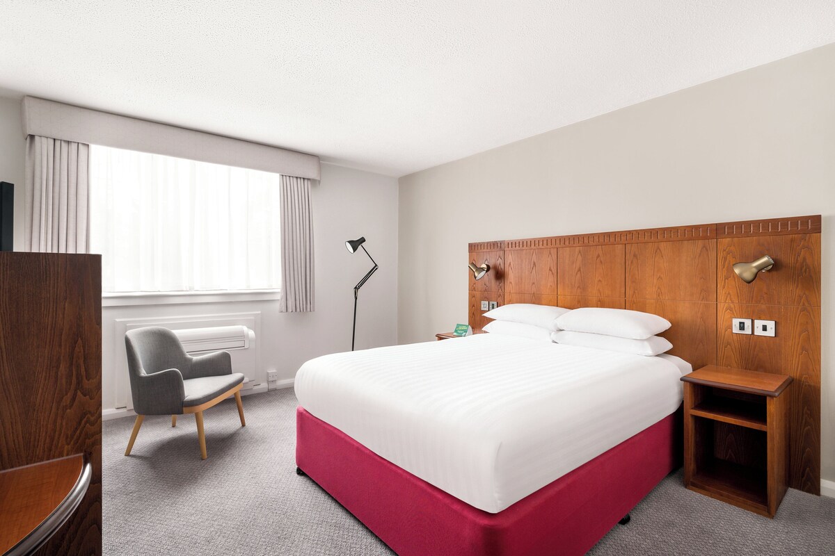 Habitación de hotel con una cama grande, un escritorio, una silla, una mesita de noche, una lámpara y una ventana con cortinas.
