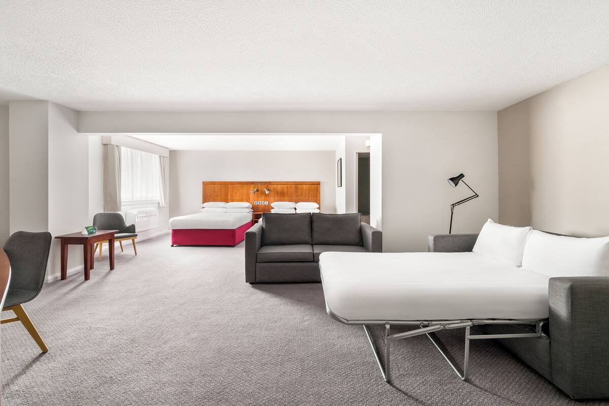 Una habitación de hotel moderna con un sofá gris, una cama blanca, un taburete rojo, un cabecero de madera y un escritorio con una lámpara.