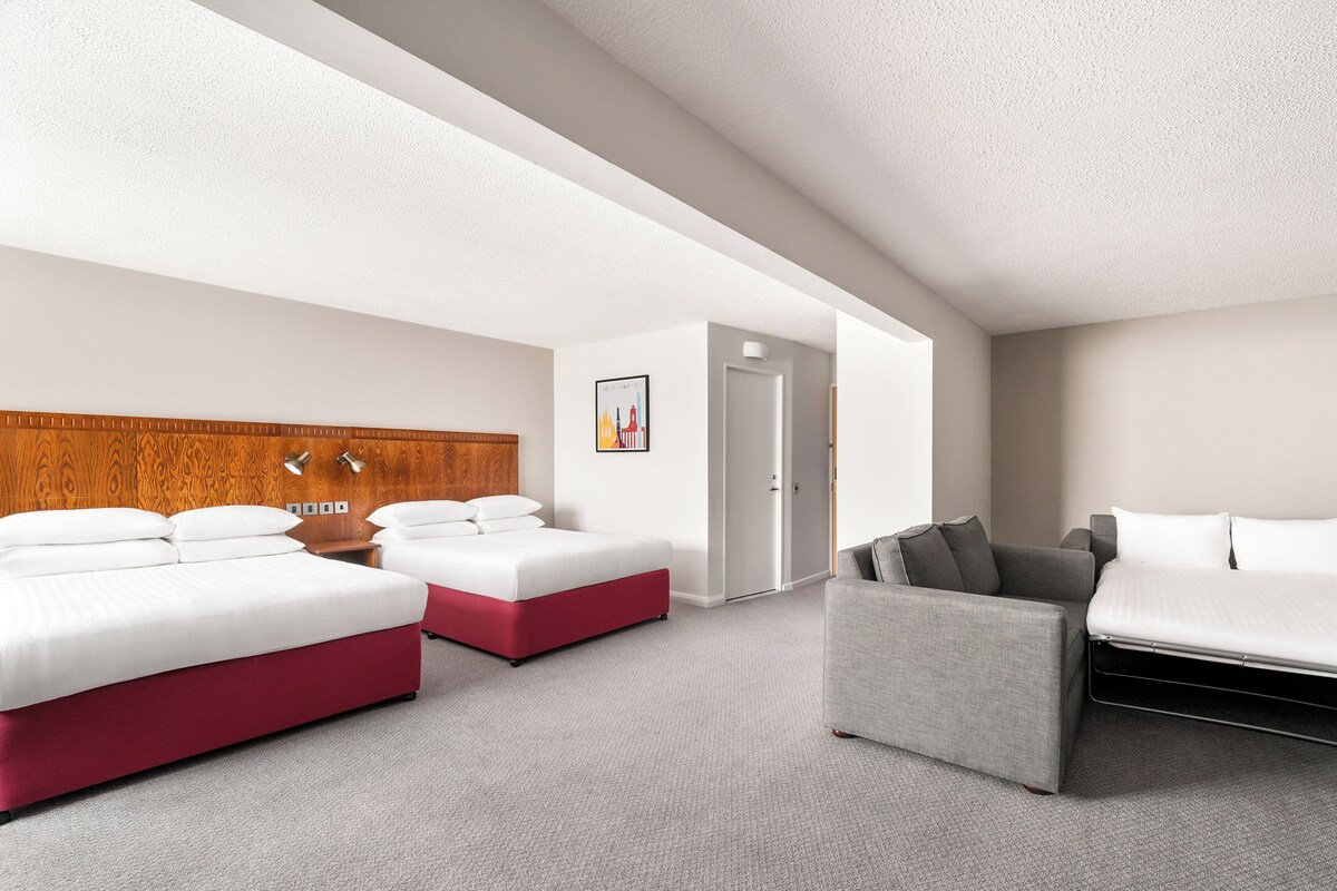Una habitación de hotel moderna con dos camas, un sofá gris y un cabecero de madera.