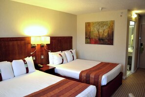 A hotel room with two beds, a nightstand with a lamp, a painting on the wall, and a bathroom door visible.