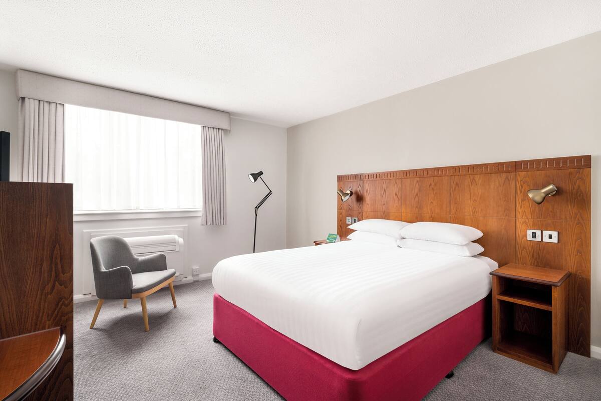 Habitación de hotel con una cama grande, un escritorio, una silla, una mesita de noche, una lámpara y una ventana con cortinas.