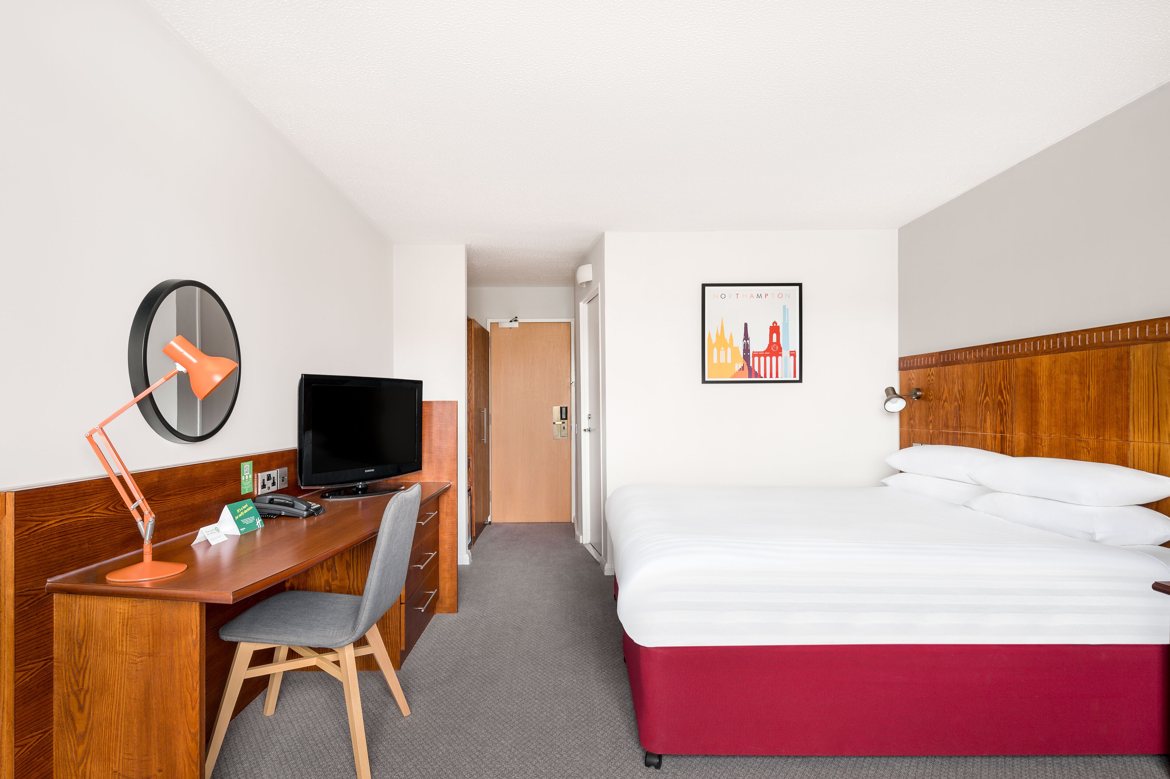 A hotel room with a wooden desk, a computer, a TV, a bed with white and red bedding, a framed picture on the wall, and a door leading to another room.