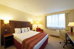 A hotel room with a large bed, a desk, a chair, a nightstand, and a window with curtains.