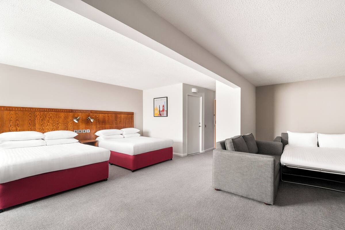 Una habitación de hotel moderna con dos camas, un sofá gris y un cabecero de madera.