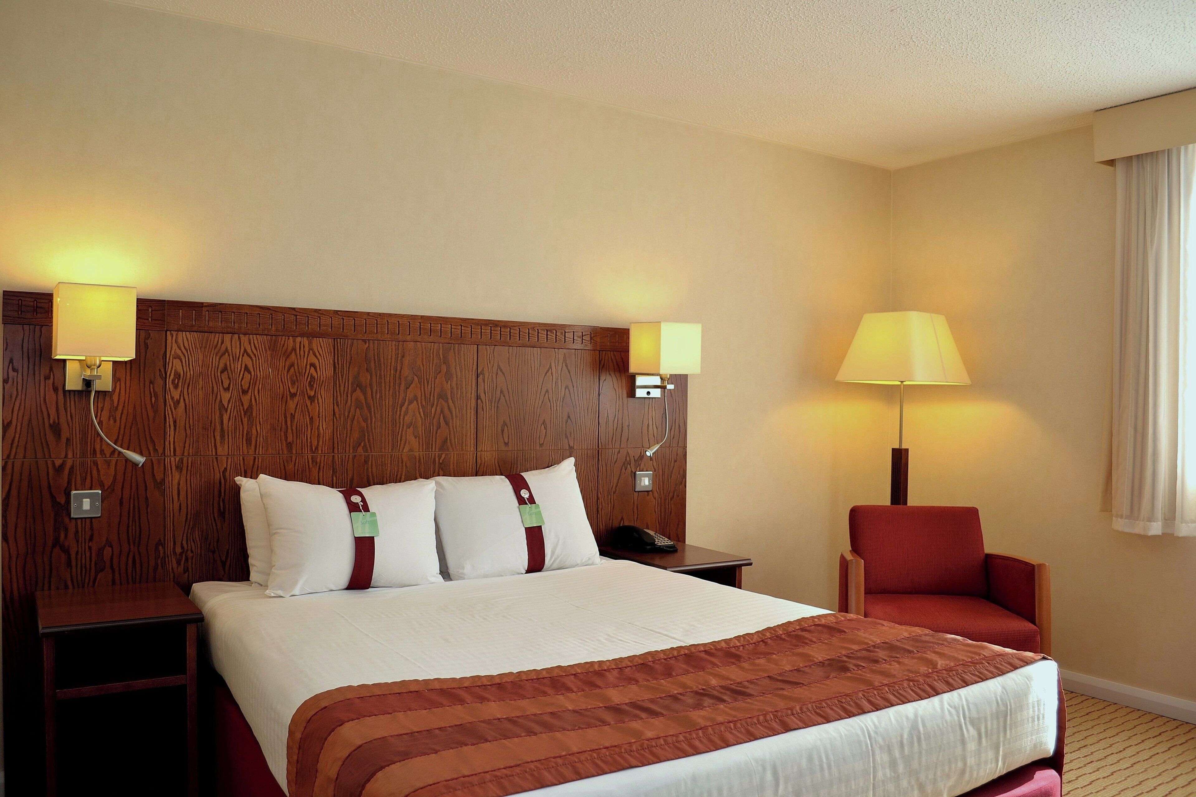 Una habitación de hotel con una cama grande, dos mesitas de noche, un sillón rojo y un cabecero de madera.
