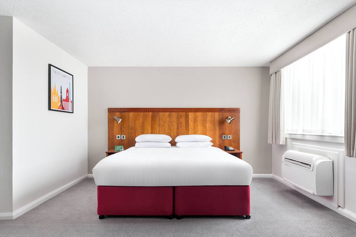 Una habitación de hotel con una cama grande, un cabecero de madera, aire acondicionado de pared y una ventana con cortinas.