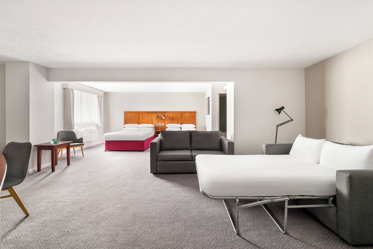 Una habitación de hotel moderna con un sofá gris, una cama blanca, un taburete rojo, un cabecero de madera y un escritorio con una lámpara.