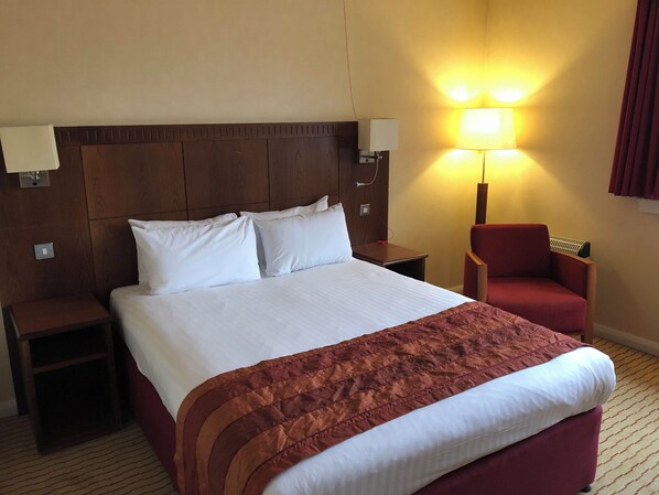 Quarto de hotel com uma cama grande, uma poltrona vermelha, uma mesa de cabeceira e um abajur.