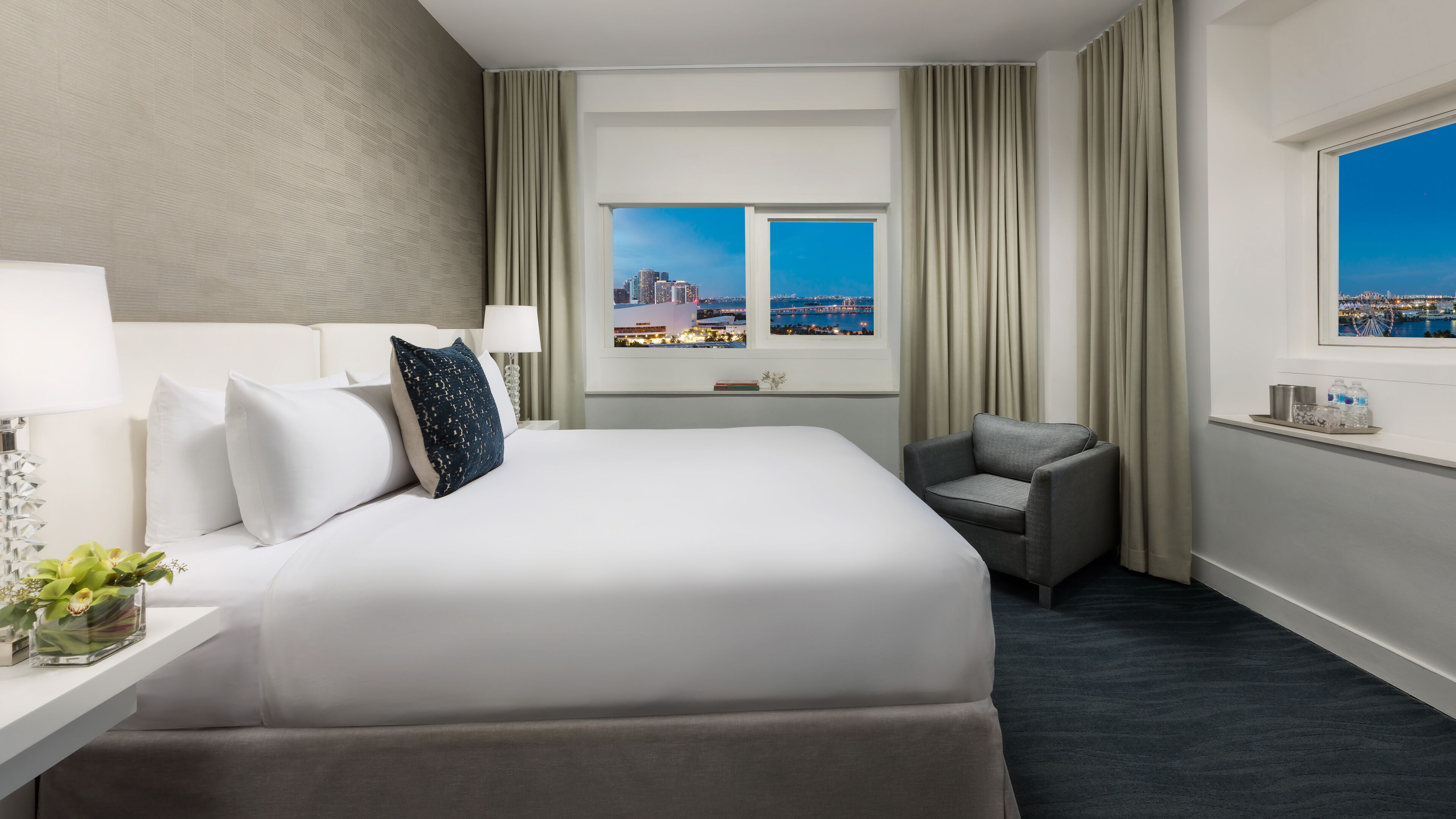 A hotel room with a large bed, a chair, a nightstand with a lamp, a small table with a vase of flowers, and a window with a city view.