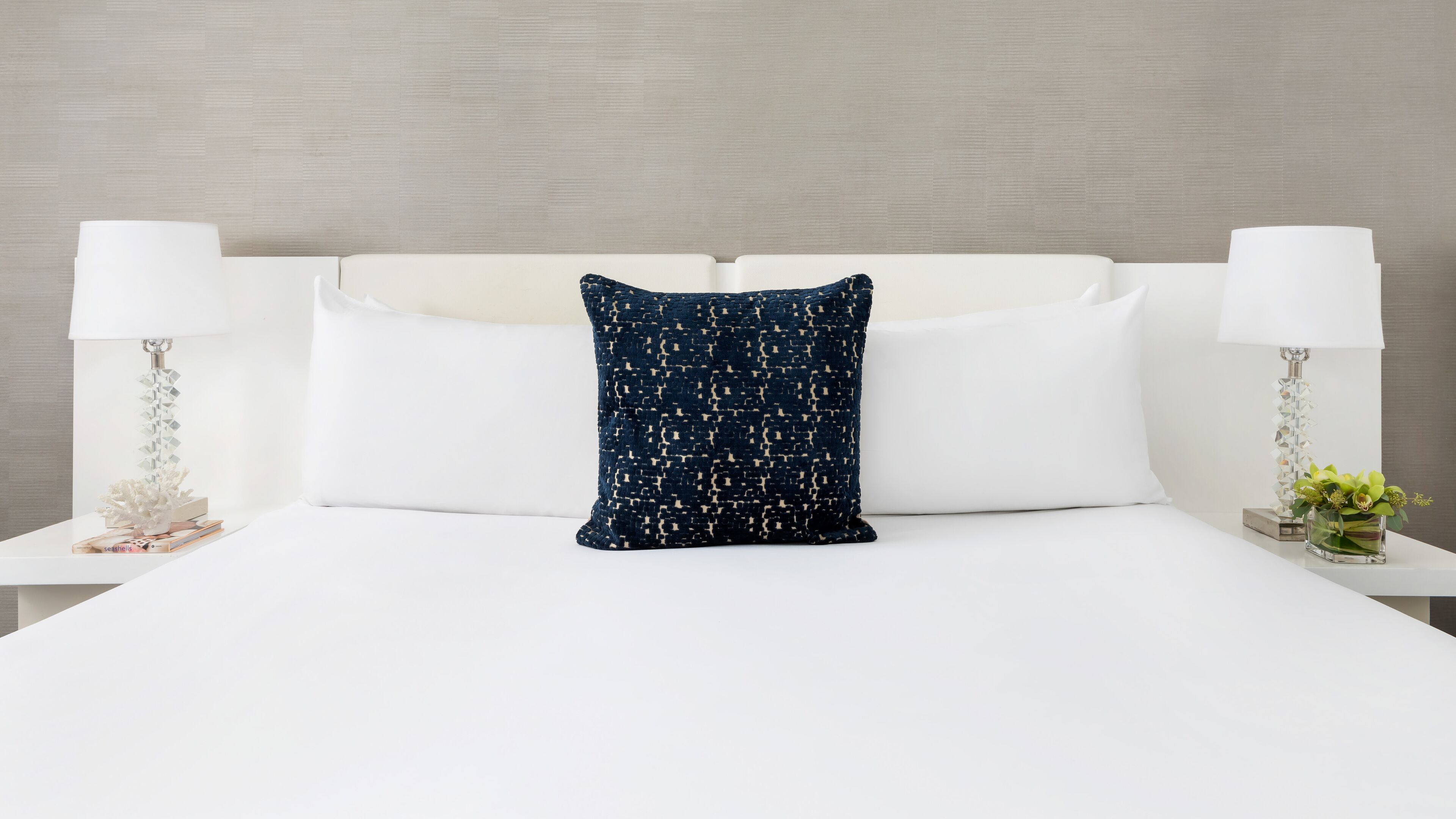 A neatly made bed with white linens, a single decorative pillow, and two bedside tables with lamps and small decorative items.