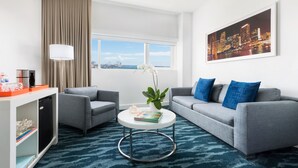 A modern hotel room with a grey sofa, a small round coffee table, a bookshelf, and a large window with a cityscape view. - YVE Hotel Miami (Miami)