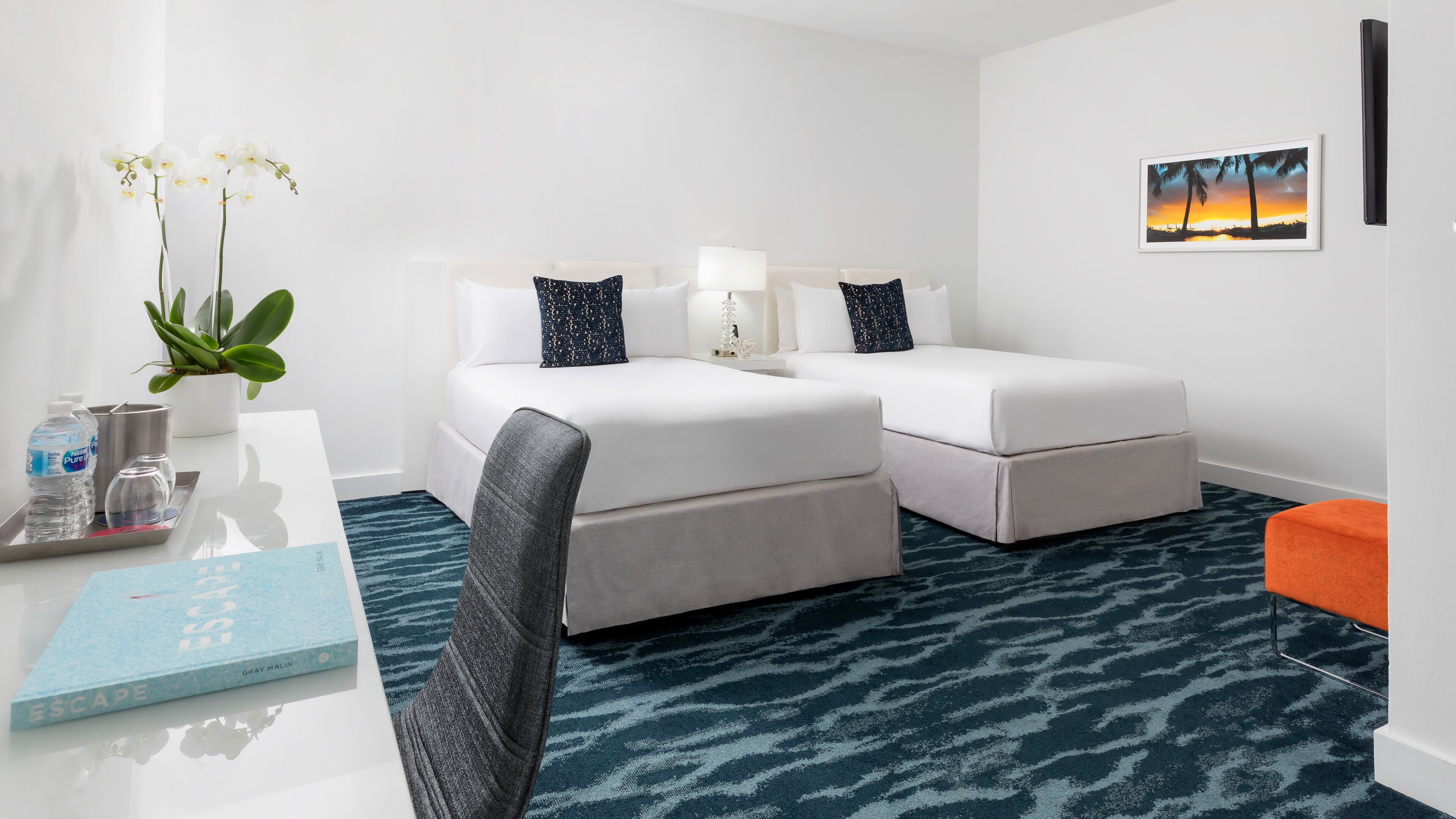 Quarto de hotel com cama, escrivaninha com um livro, planta e um quadro na parede.