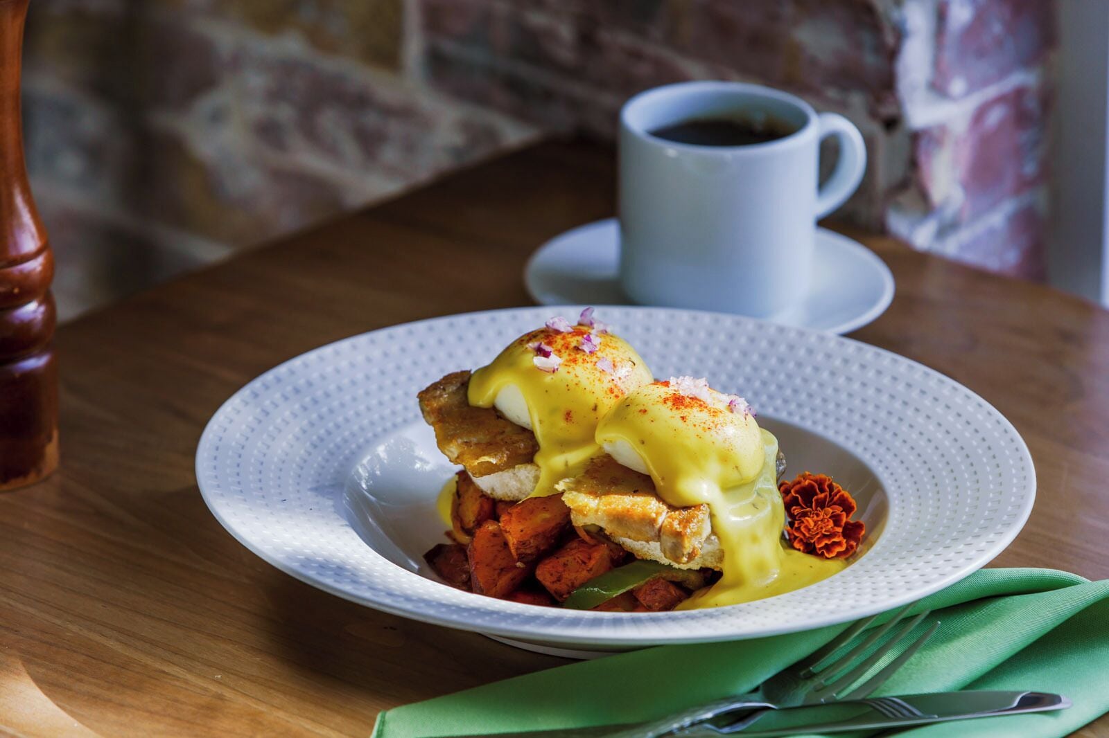 A plate of food with eggs benedict and hash browns, a cup of coffee, and a green napkin on a wooden table.