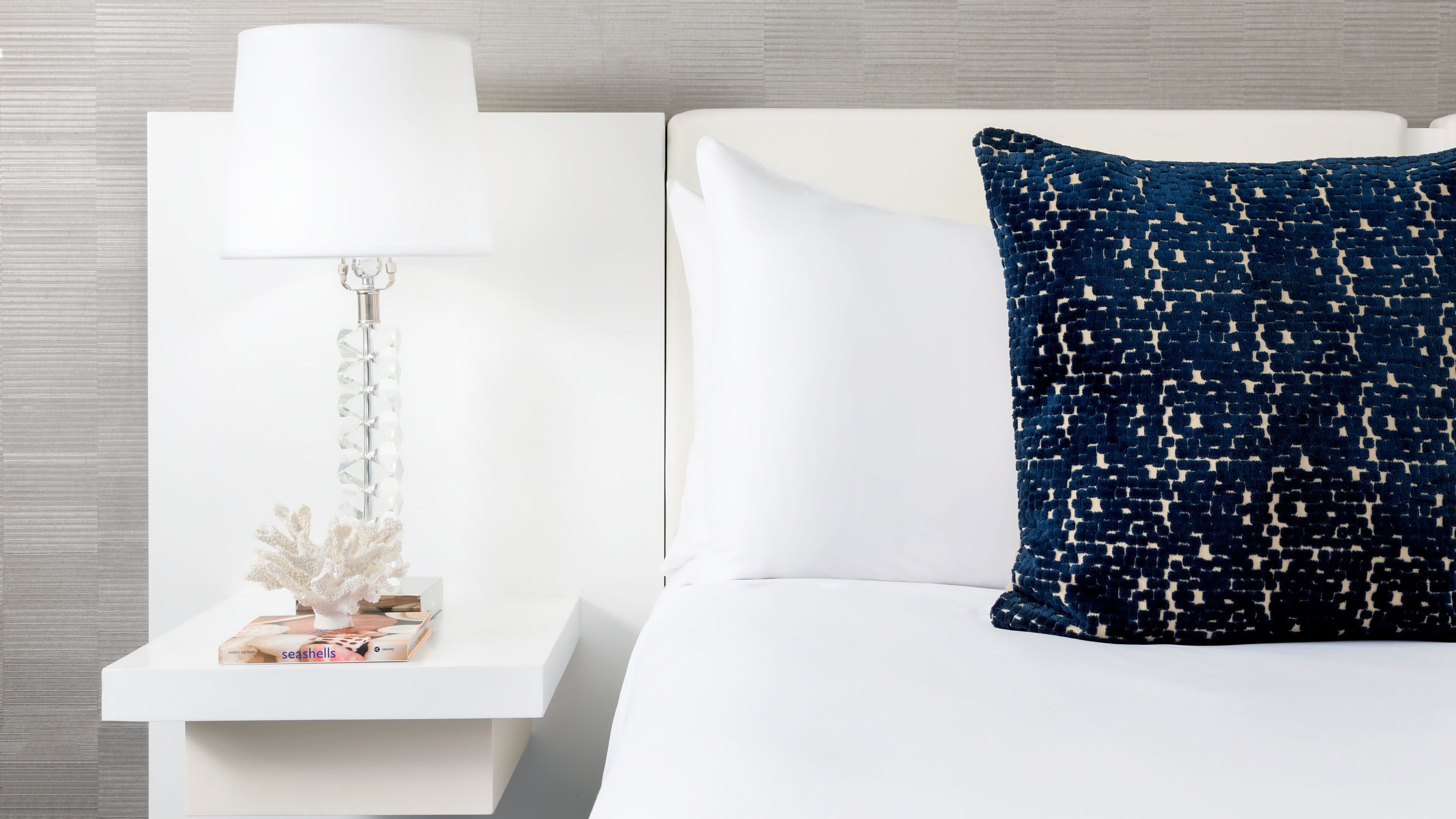 A neatly made bed with a white headboard, a white nightstand with a lamp and decorative items, and a blue patterned pillow.
