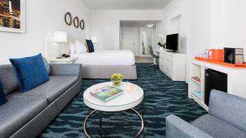 Quarto de hotel moderno com sofá cinzento, mesa de centro redonda com um livro, cama com roupa de cama branca, televisão e pequena cozinha.