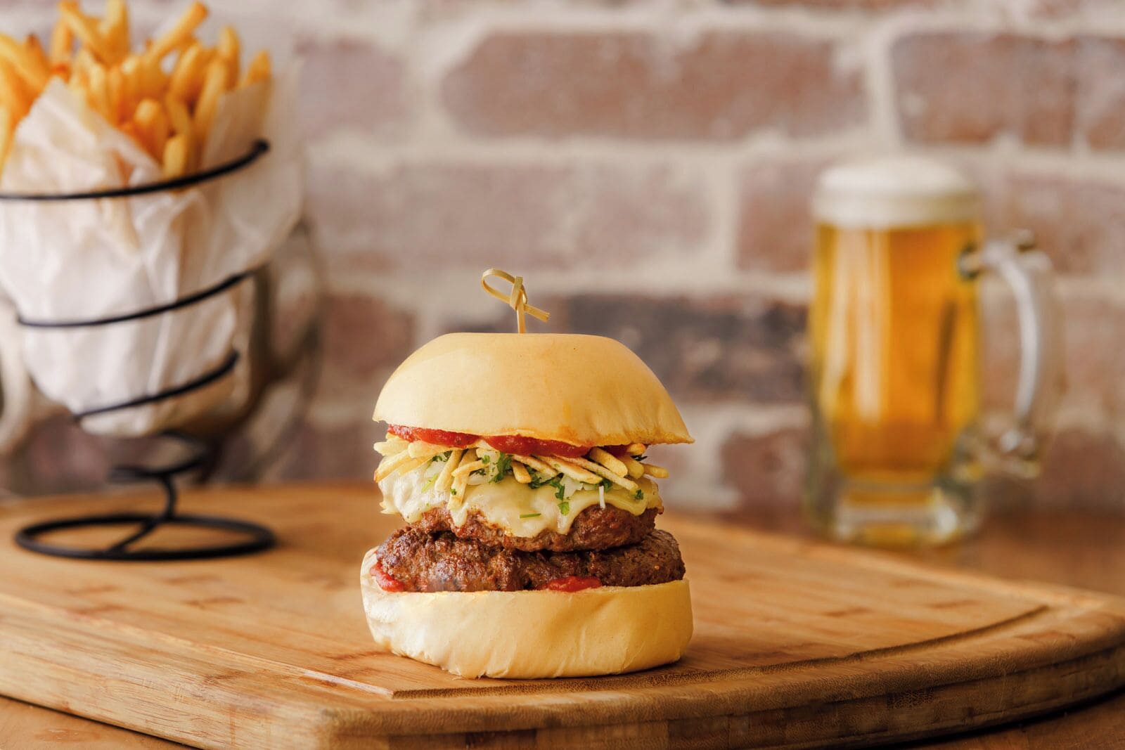 A burger with lettuce, tomato, and cheese on a bun, a glass of beer, and a basket of fries on a wooden board.