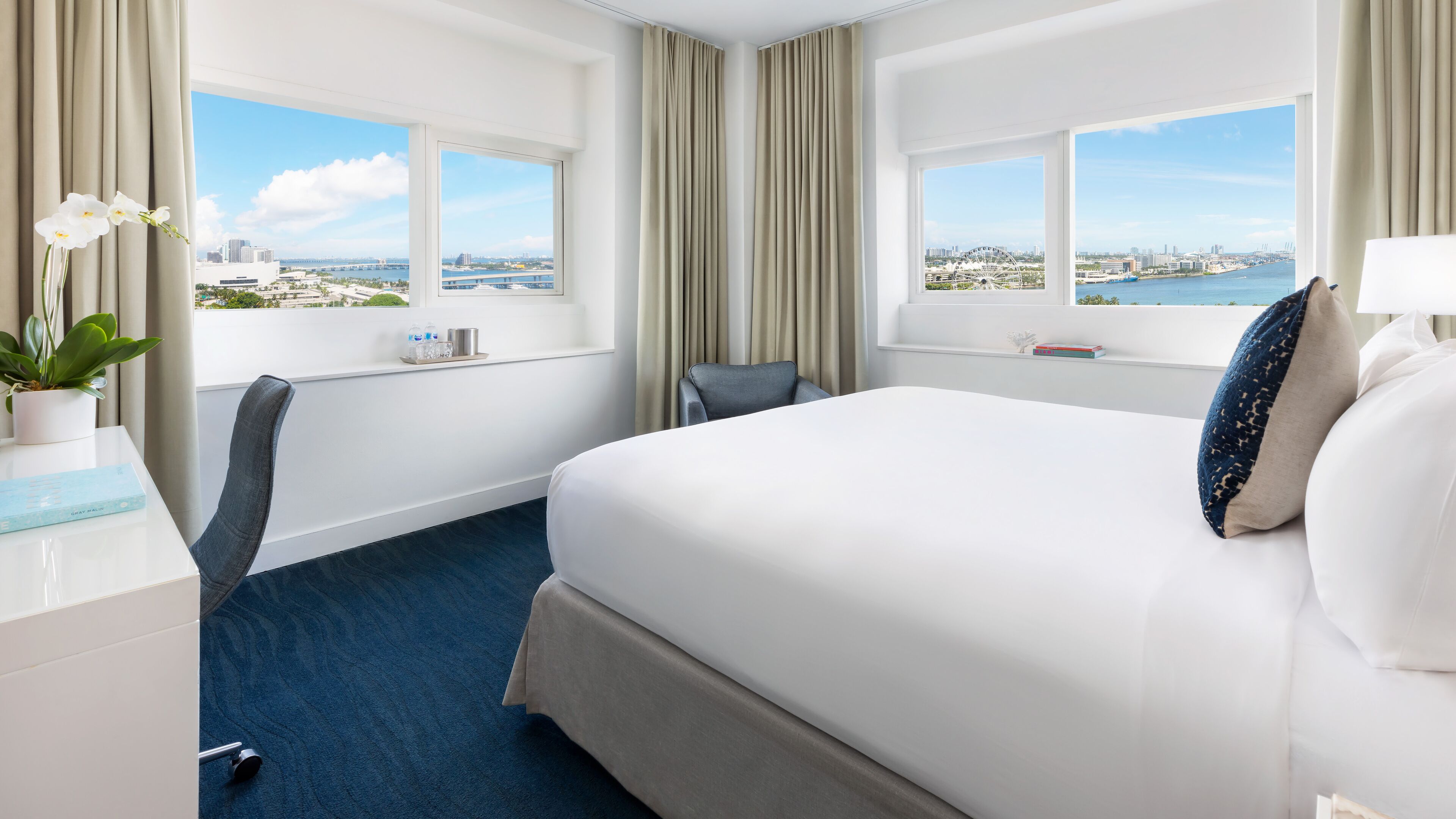 A hotel room with a large bed, a desk, a chair, and a view of the city and water.