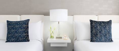Une chambre d’hôtel avec deux lits blancs, chacun agrémenté d’un oreiller à motifs bleu marine, une table de chevet blanche avec une lampe et un vase rempli de fleurs.