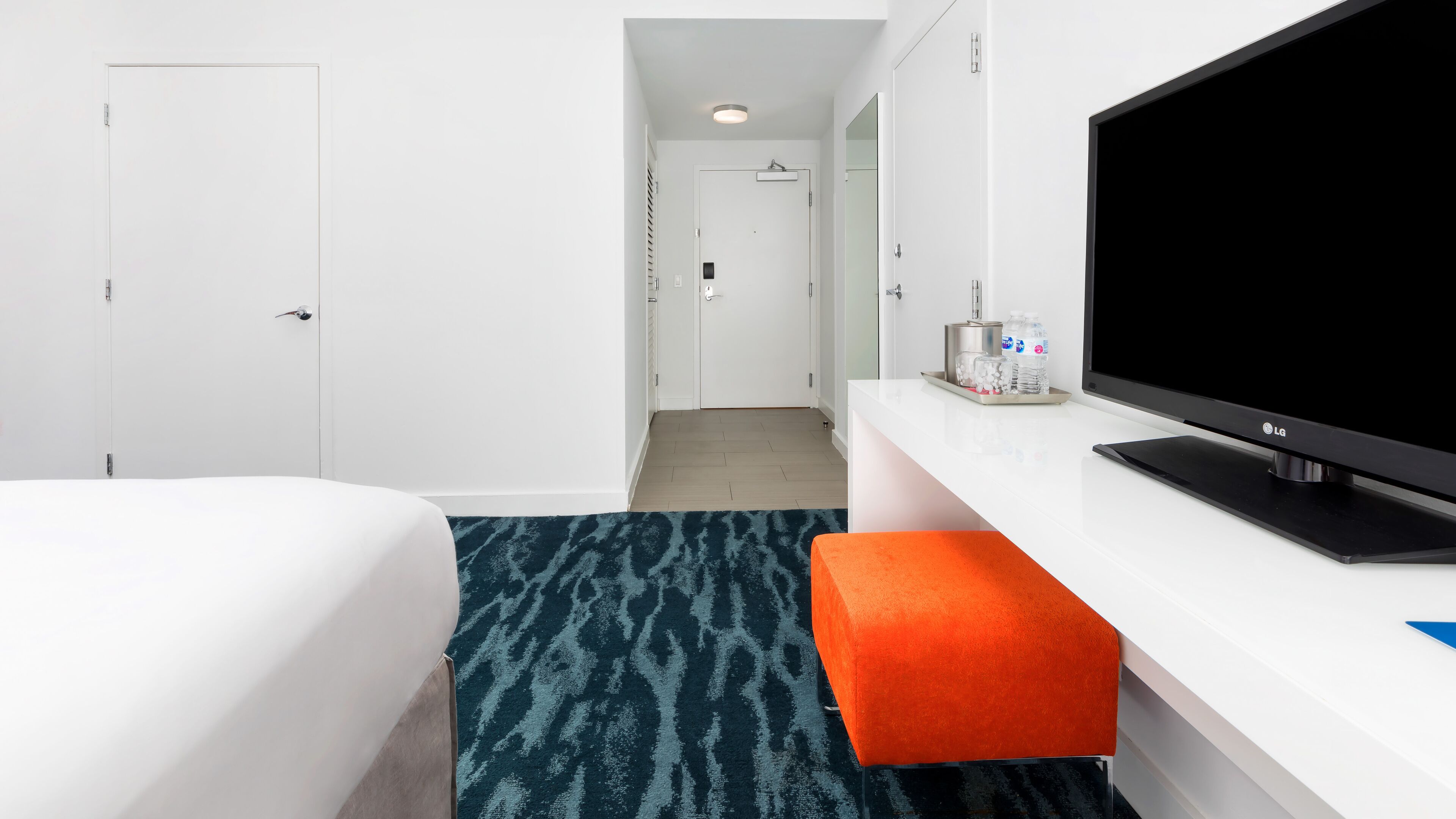 Habitación de hotel con cama, televisión, una mesita con una botella de agua y una puerta de baño al fondo.