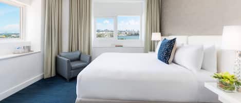 A hotel room with a large bed, a chair, a desk with a lamp, and a view of a cityscape through the window.