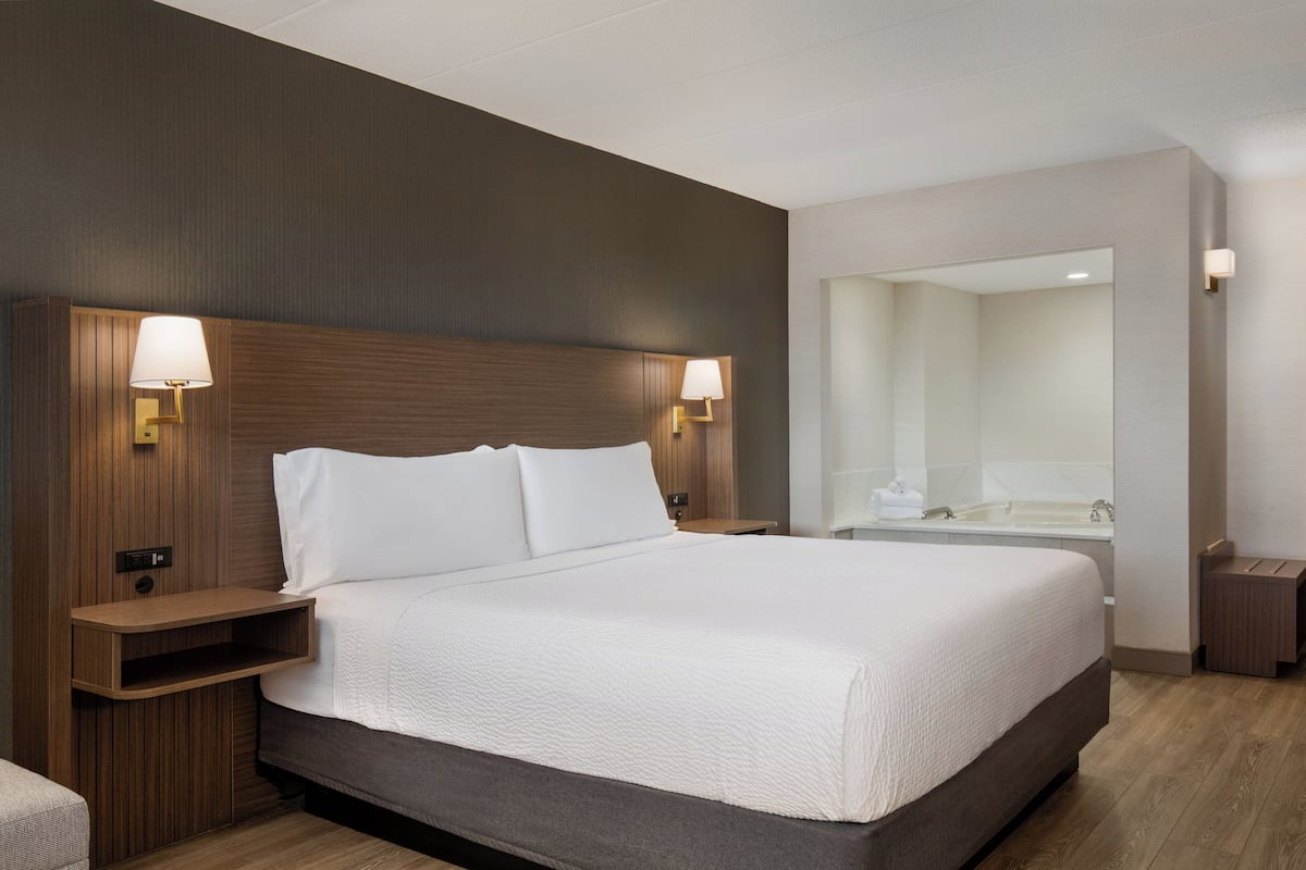Habitación de hotel moderna con una cama grande, cabecera de madera, lámparas de noche, mesitas de noche y un baño visible a través de una puerta abierta.