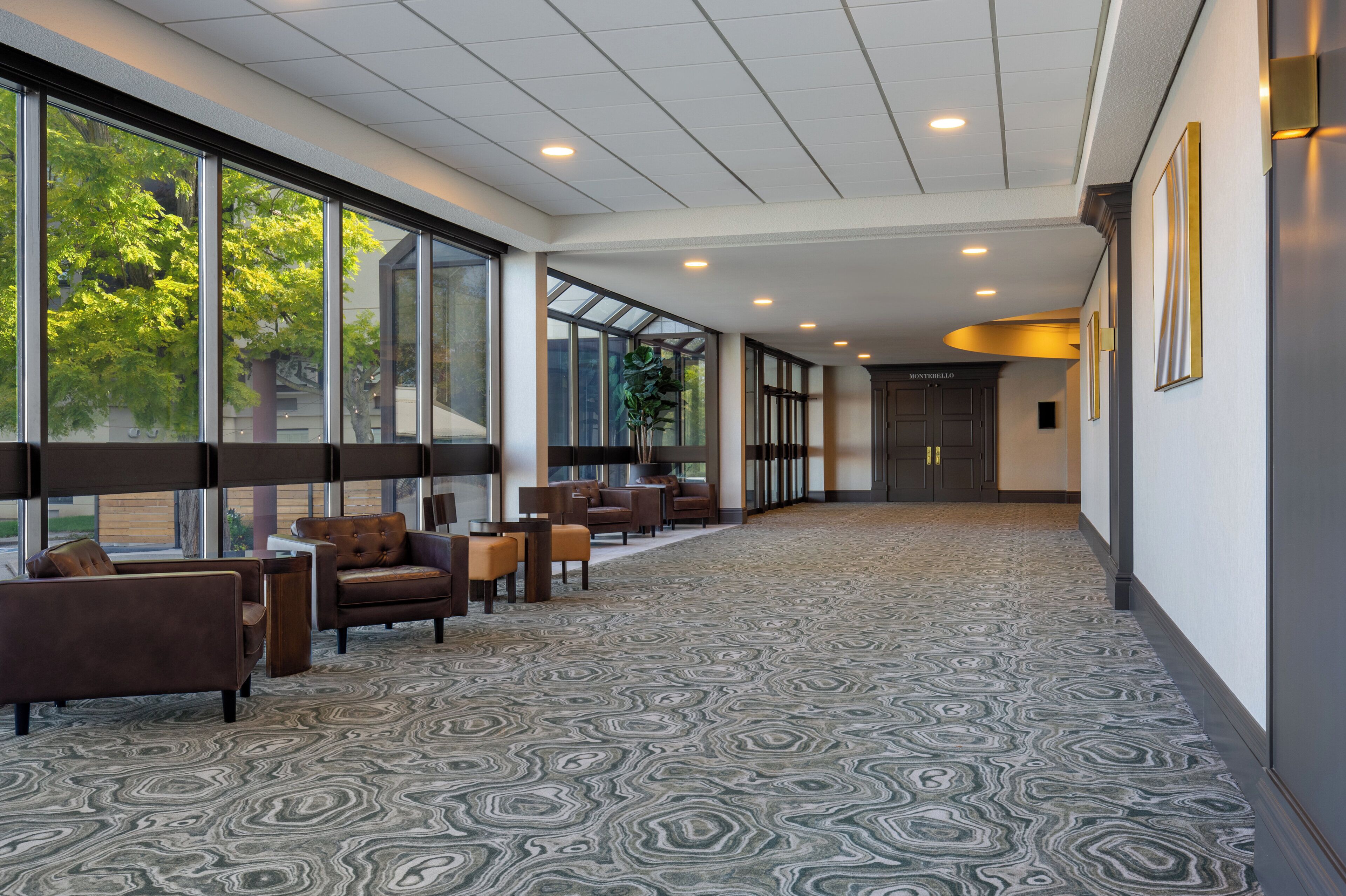 A spacious hallway with a patterned carpet, large windows offering views of trees, and a seating area with leather chairs.