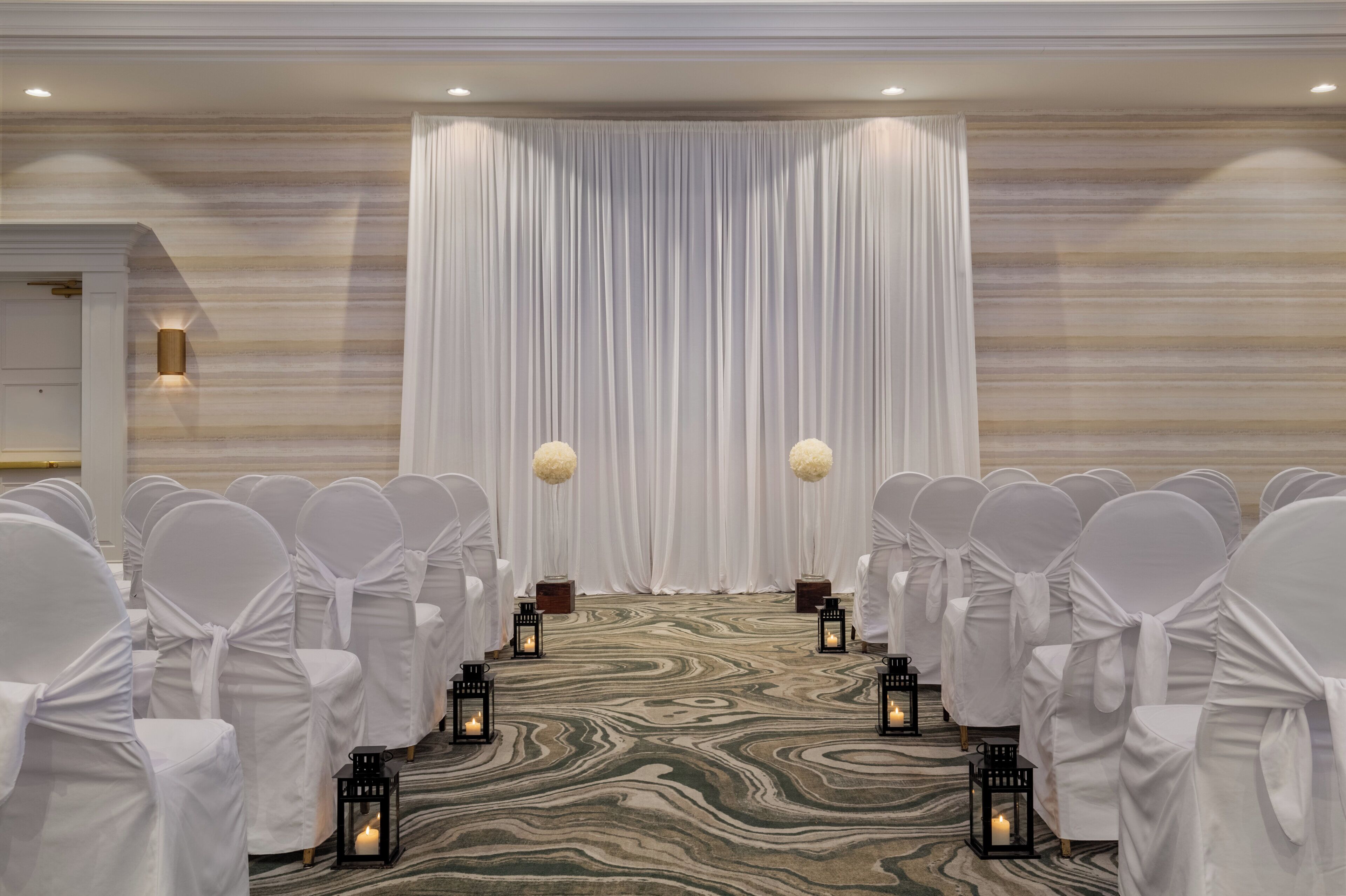 A formal event space with rows of white-covered chairs, a draped backdrop, and lanterns on a patterned carpet.