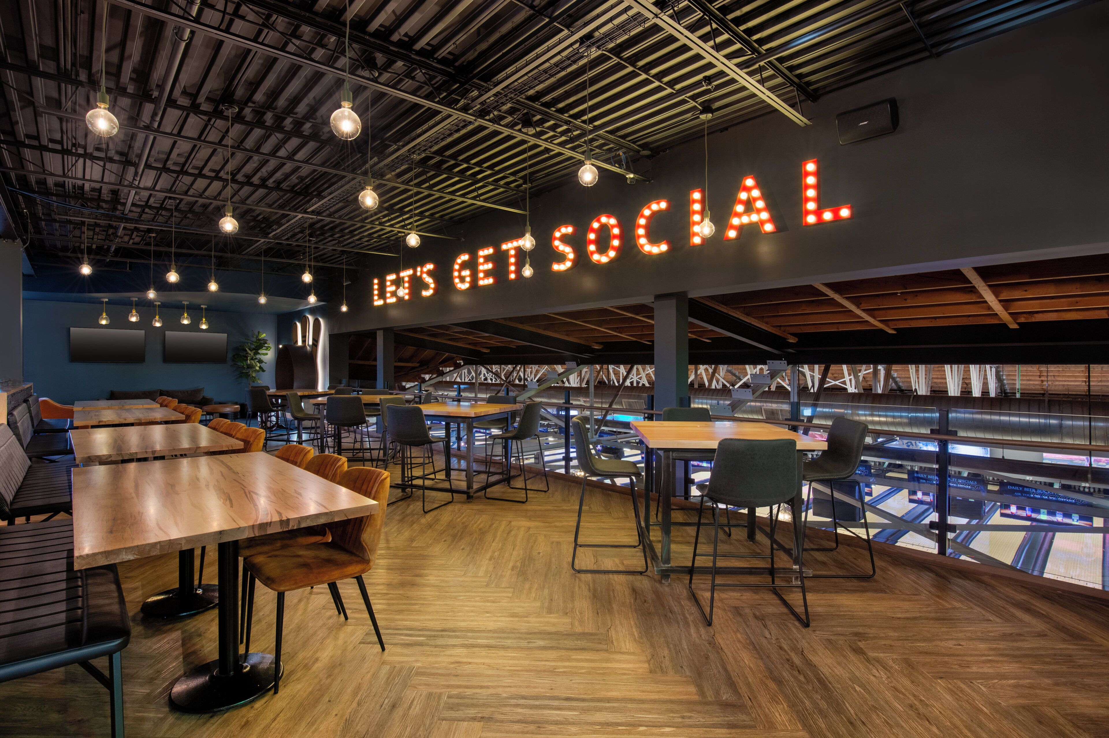 A modern cafe with wooden tables and chairs, a large illuminated sign reading "LET'S GET SOCIAL," and a view of a sports stadium through large windows.