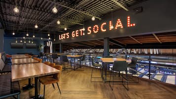 A modern cafe with wooden tables and chairs, a large illuminated sign reading "LET'S GET SOCIAL," and a view of a sports stadium through large windows.