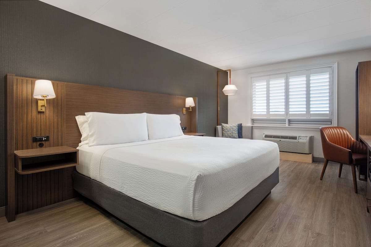 Una habitación de hotel moderna con una cama grande, cabecero de madera, lámparas de noche, un escritorio y una ventana con persianas.