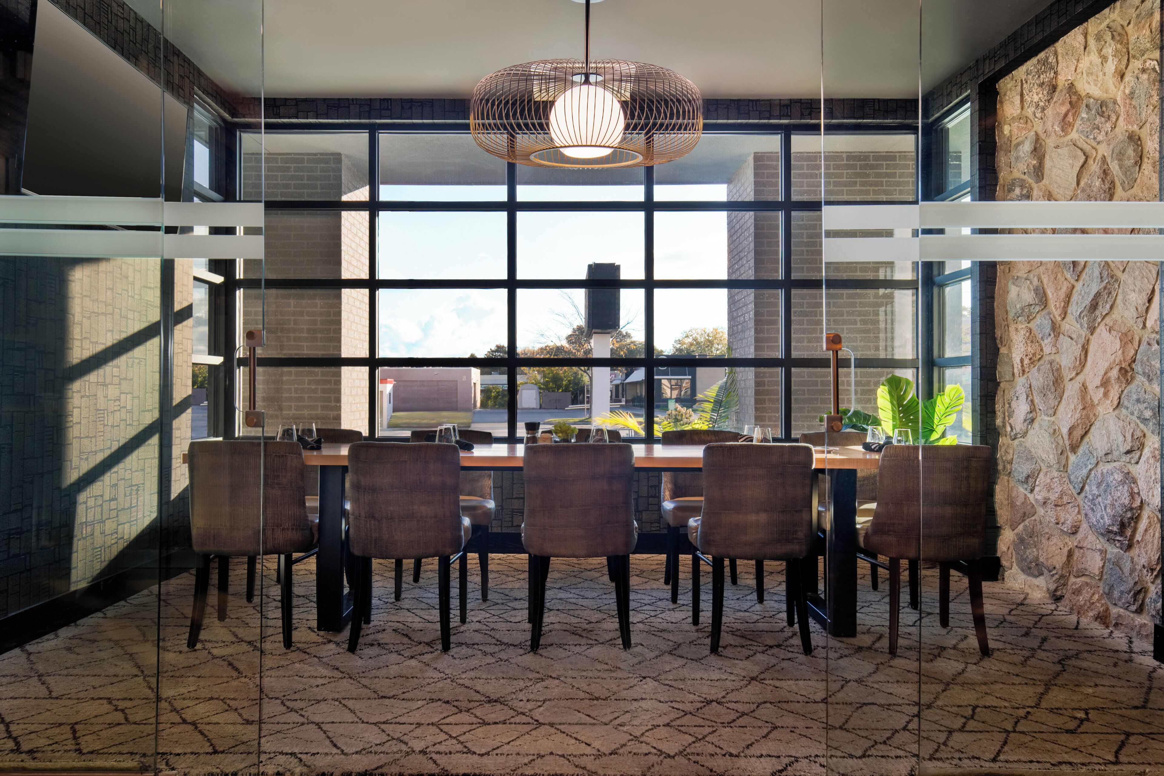 A modern dining area with a large table and chairs, large windows, and a stone wall.