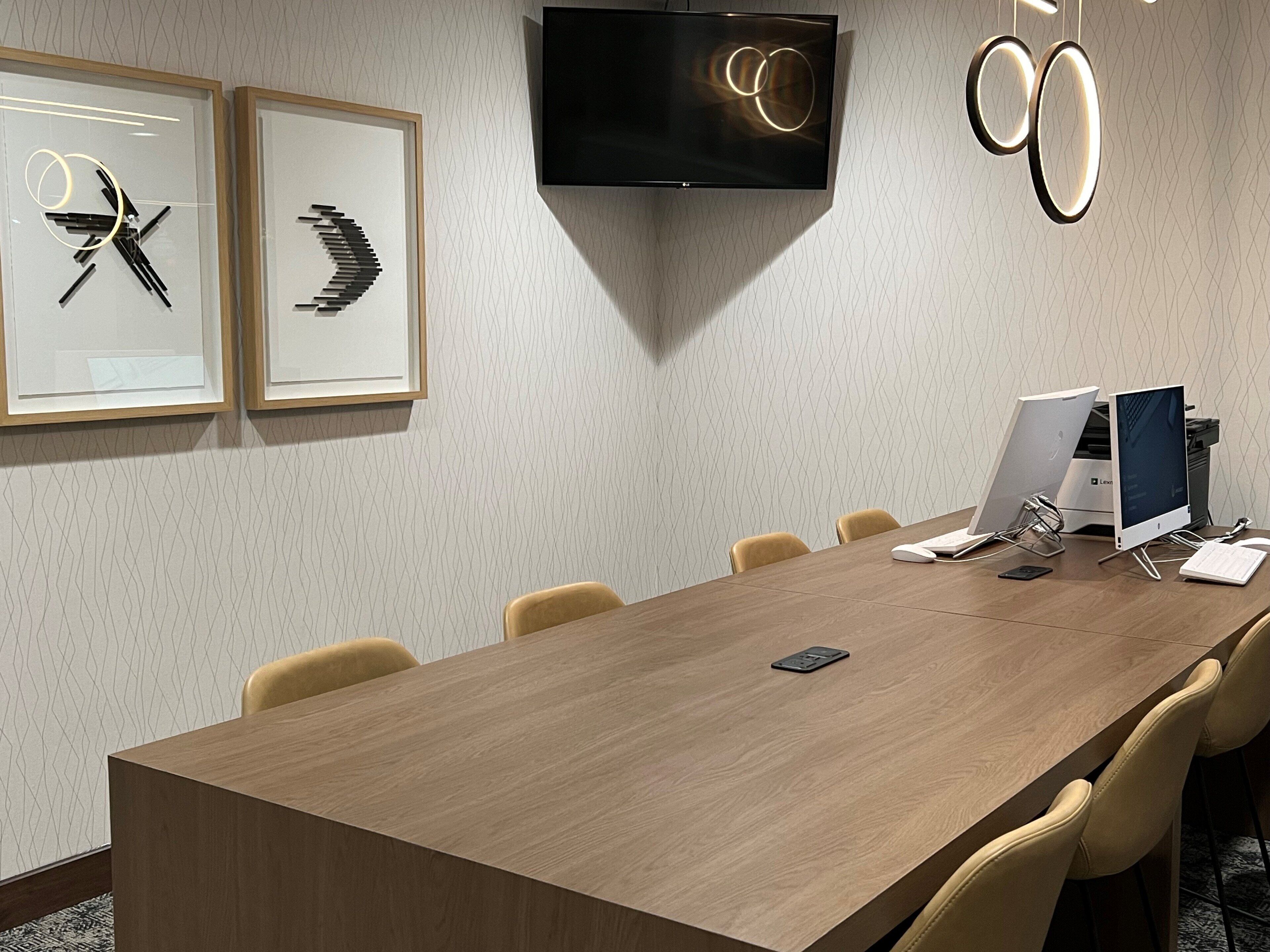 A conference room with a long wooden table, six chairs, a flat-screen TV mounted on the wall, and two framed abstract artworks.