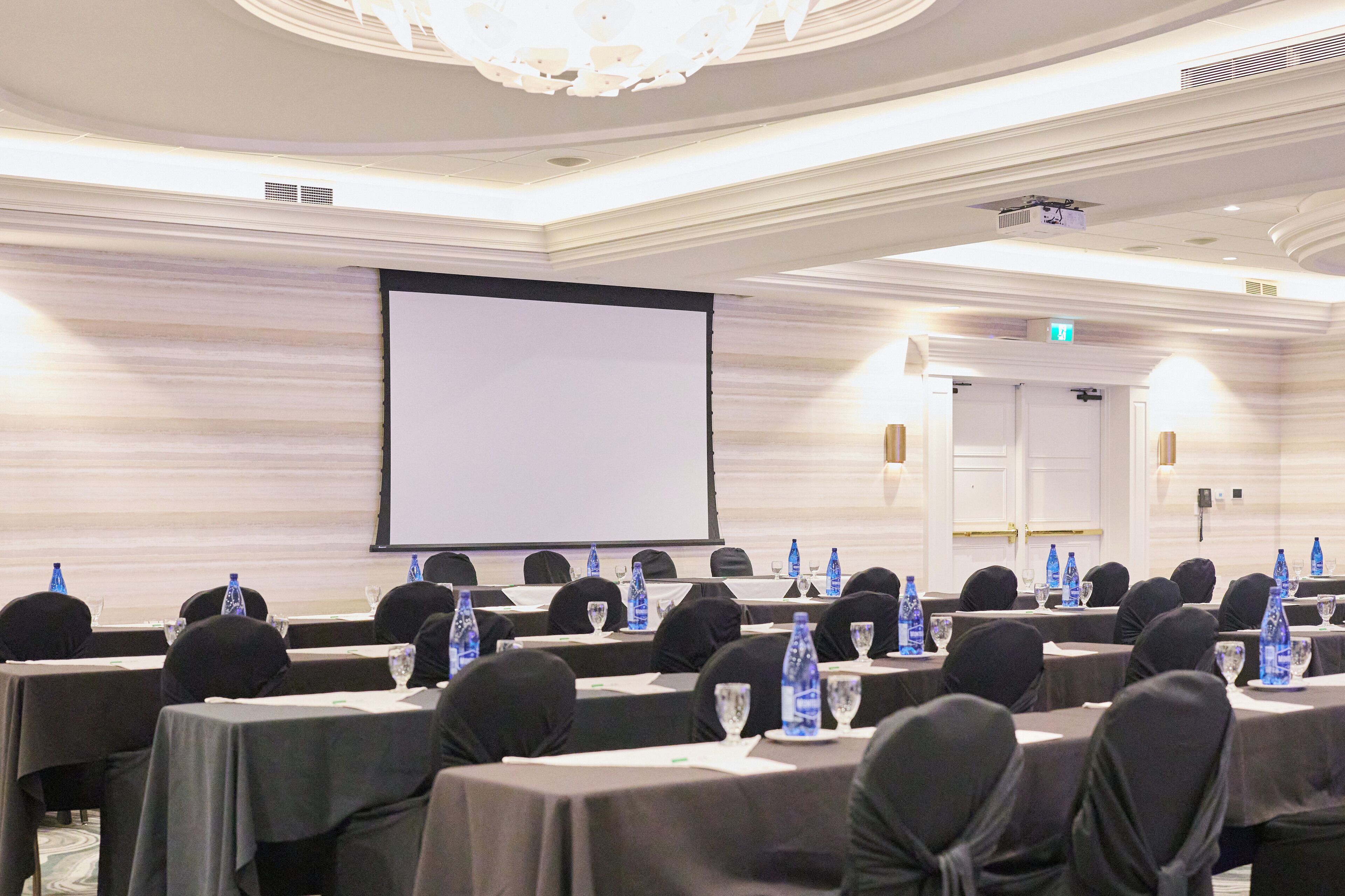 A conference room with tables set for a meeting, equipped with water bottles and glasses, in a room with a large screen and elegant ceiling design.