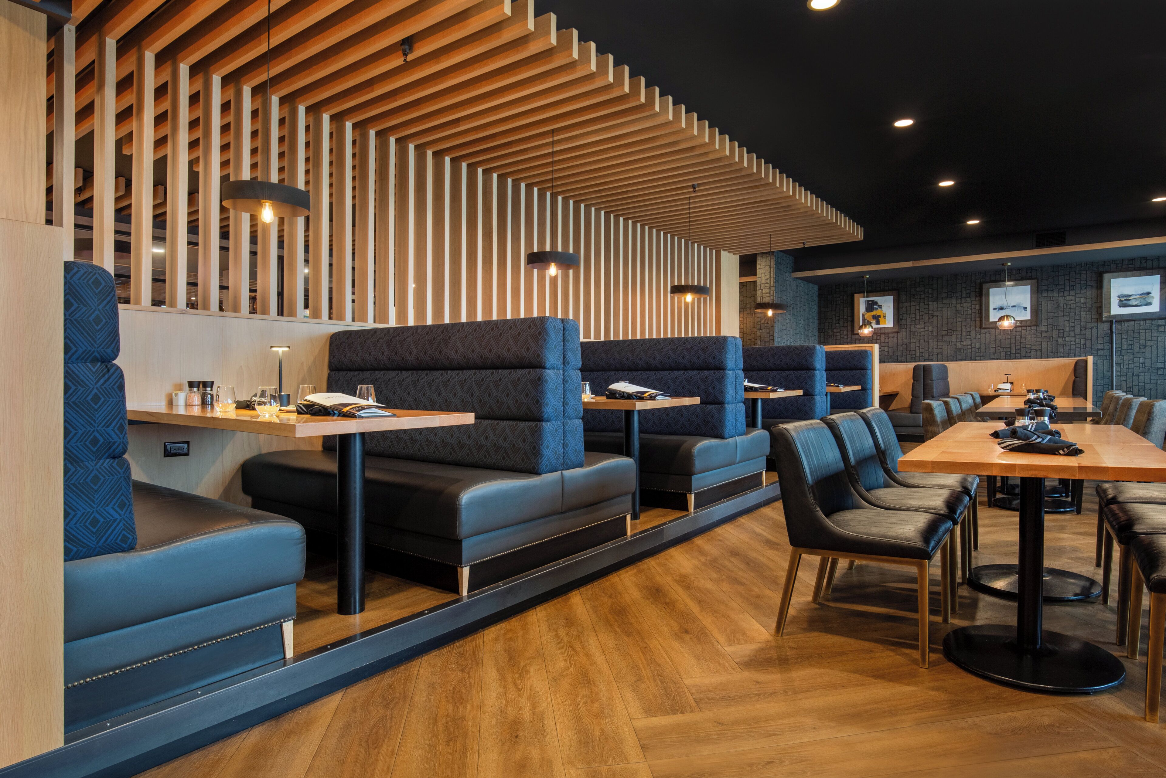 A modern restaurant with wooden paneling, blue cushioned booths, and black upholstered chairs.